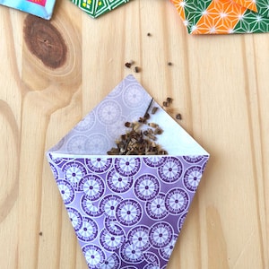 May include: A folded paper seed packet featuring a purple and white circular geometric pattern, containing a small pile of brown seeds. The packet is open at the top, revealing the seeds inside, with other colorful patterned paper packets visible nearby.