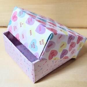 May include: A rectangular paper box with a lid featuring a pattern of pastel-colored hearts printed with phrases like "LOVE," "HUGS," "HOT STUFF," "BEST DAY," "CUTIE PIE," "XOXO," "SAY YES," "4 EVER," and "SOUL MATE." The base has a light pink arrow pattern.