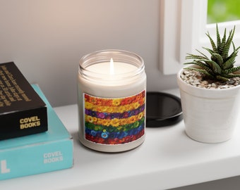May include: A white candle with a rainbow floral design on the label. The candle is lit and the flame is visible. The candle is sitting on a white shelf with books and a potted plant.