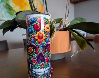 May include: A white, stainless steel tumbler with a colourful floral skull design. The artwork features flowers in red, orange, yellow, blue, and purple. The cylindrical tumbler has a silver lid.