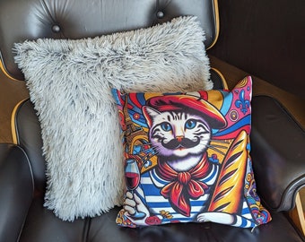 May include: A colourful cushion with a cat wearing a beret and scarf, holding wine and a baguette. A fluffy grey cushion sits behind it on a black leather chair.
