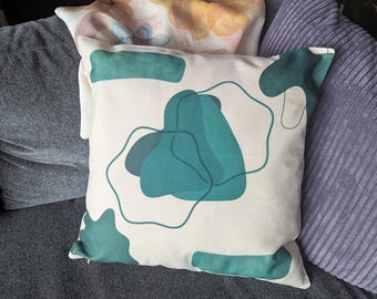 May include: Square cushion with a cream background and teal abstract shapes. The cushion is on a grey sofa next to a purple corduroy cushion and a floral cushion.
