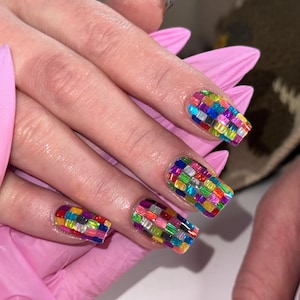 May include: Close-up of manicured nails with a colorful, mosaic-like design. Each nail features a rectangular shape with a pattern of small, iridescent, multi-colored rectangles. The nails are set against a soft pink background.