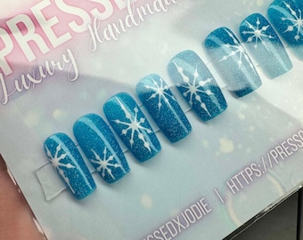 May include: A set of blue glitter press-on nails with white snowflake designs. The nails have a gradient effect, transitioning from a darker blue to a lighter shade. The card reads "PRESSED BY JODIE" and the Instagram handle @PRESSEDXJODIE.