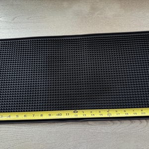 May include: Black rectangular bar mat with a textured surface. A yellow measuring tape shows the mat is approximately 22 inches long. The mat is designed to catch spills and protect surfaces.
