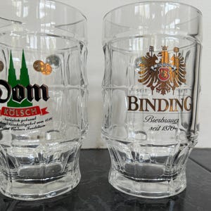 May include: Two clear glass beer mugs. One mug features the word "Dom" with a green and gold design, and the other mug has the word "Binding" with a gold emblem. Both mugs have a square shape and a thick base.