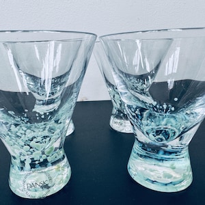 May include: Four clear martini glasses with a unique design. The base and lower portion of the glasses feature a speckled, seafoam green and blue pattern. The glasses are made by Dansk and are recommended to be hand washed.