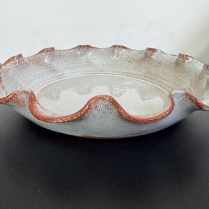 May include: A handmade ceramic pie dish with a ruffled edge. The dish has a light gray interior and a reddish-brown rim. The pie plate is round and has a rustic, textured appearance.