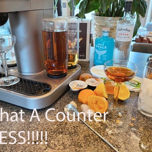 May include: A bar setup with a cocktail machine, glassware, and various ingredients. Includes bottles of vodka and gin, along with sliced oranges, honey, and ice. The text "What A Counter MESS!!!!" is visible.