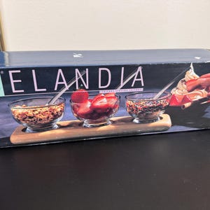 May include: A Selandia condiment set in a box. The set includes three clear glass bowls with spoons, set on a wooden tray. The bowls contain granola, strawberries, and sprinkles. The box has the words "Condiment Set" and "Selandia" printed on it.