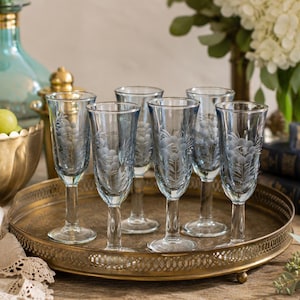 May include: Six clear champagne flutes with a blue tint and floral etched design are arranged on a gold-toned metal tray. A gold-colored bowl with green grapes and a decorative glass bottle are in the background.