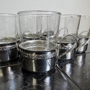 May include: Set of six clear glass tumblers with silver-colored metal holders. The holders feature an ornate, scrolled design around the base and a handle. The glasses sit on a dark, reflective surface.