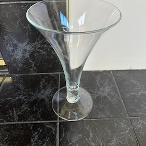 May include: A clear, stemmed glass with a wide, flared bowl. The glass has a slender stem and a circular base. The glass is designed for serving cocktails or other beverages. The glass is made of transparent glass.