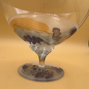 May include: A clear glass goblet with a wide bowl and a frosted base. The bowl features a painted landscape scene with brown and purple bison. The stem is slender and the base is round.