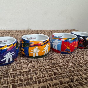 May include: Four Skye Mcghie fine porcelain bowls with colorful abstract designs. Each bowl features a white interior and a unique exterior pattern with blues, yellows, reds, and white figures. The bowls are arranged on a textured brown surface.