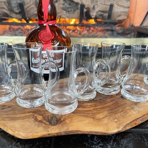 May include: A collection of clear glass cordial glasses with handles, arranged on a wooden serving board. A bottle of Grand Marnier liqueur is in the background. The glasses are in front of a fireplace.