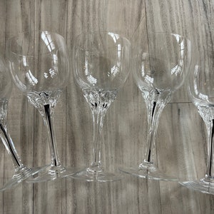 May include: Five clear wine glasses with a decorative base and a black stem. The glasses are arranged on a light-colored wooden surface. The glasses are empty and appear to be made of crystal.
