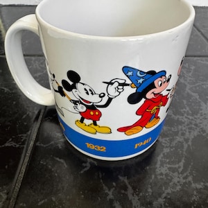 May include: White ceramic mug featuring Mickey Mouse in various poses, including a wizard. The mug has a blue band at the bottom with the years 1932 and 1940. The handle is white and the interior is also white.