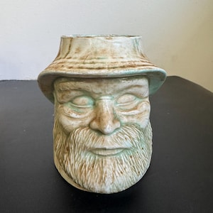 May include: A ceramic mug shaped like the head of a person wearing a wide-brimmed hat. The mug features a sculpted face with closed eyes, a prominent nose, and a textured beard. The finish is a pale green glaze with brown accents in the crevices.