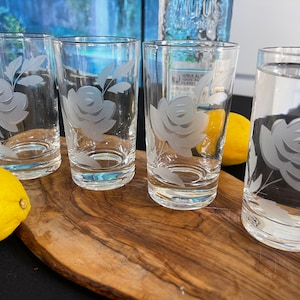 May include: Four clear glass tumblers with etched rose designs. The glasses are arranged on a wooden board with two yellow lemons. The glasses are filled with water. The background includes a blue and white painting.