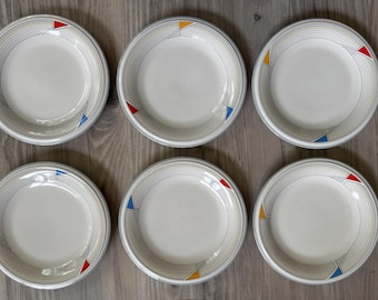 May include: Six white ceramic bowls with a gray rim and concentric gray lines. Each bowl has a colorful geometric design with red, yellow, and blue triangles. The bowls are arranged on a wooden surface.