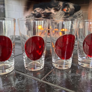 May include: Four clear drinking glasses with a large, red, oval-shaped design on the side. The glasses are cylindrical and appear to be made of glass. The background shows a fireplace.