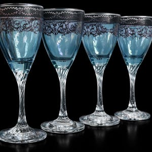 May include: Four elegant, vintage-style wine glasses with a light blue hue. Each glass features a silver rim with a black floral design and a twisted stem. The glasses are set against a black background.