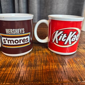 May include: Two ceramic mugs on a wooden surface. One mug is dark brown with "Hershey's s'mores" printed on it. The other mug is red with the "Kit Kat" logo. Both mugs have white handles and rims.