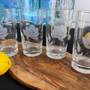 May include: Four clear glass tumblers with etched rose designs. The glasses are cylindrical and sit on a wooden board, with a yellow lemon to the left. The background is blurred, suggesting a kitchen or dining setting.