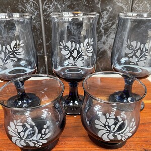 May include: Set of five blue-tinted glass goblets and tumblers. Each glass features a white floral design. The goblets have a stem and the tumblers have a short base. The glasses are arranged on a wooden surface.