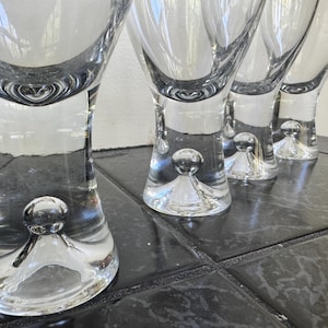 May include: Four clear, stemmed wine glasses with a unique design. Each glass features a bulbous base with a small, spherical detail. The glasses are arranged on a dark, tiled surface, reflecting light.