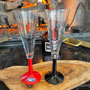 May include: Two clear champagne flutes with colored stems, one red and one black, sit on a wooden board. The red-stemmed glass has a Leonardo label. A bottle of liquor is visible in the background.