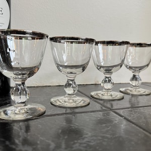 May include: Four clear glass wine glasses with silver rims. The glasses have a classic design with a rounded bowl, a decorative stem, and a circular base. The glasses are arranged in a row, showcasing their elegant shape and design.