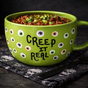 May include: A green ceramic mug filled with chili, garnished with green onions. The mug is decorated with eye-shaped designs and the phrase "Creep It Real". A black spoon and a skull-patterned napkin are nearby.