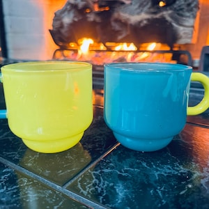 May include: Two colorful glass mugs. One is yellow with a blue handle, the other is blue with a yellow handle. The mugs are sitting on a dark, patterned surface, with a fireplace in the background.