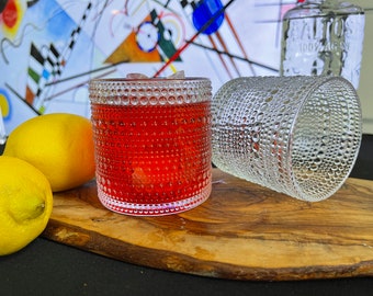 Può includere: Un cocktail rosso in un bicchiere strutturato con ghiaccio, accanto a un bicchiere trasparente con un design simile. Due limoni gialli sono su una tavola di legno. Una bottiglia di tequila è sullo sfondo.