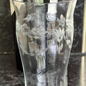 May include: A clear glass vase with etched floral and leaf designs. The vase has a flared top and a slightly curved body. The etched design is delicate and detailed, adding a touch of elegance. The background includes a dark bottle.