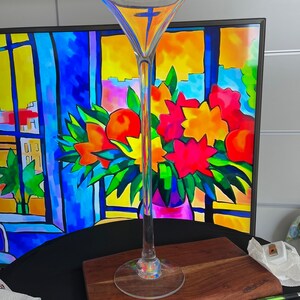 May include: A tall, clear glass martini glass with a wide bowl and long stem. The glass sits on a wooden board. The background features a colorful painting of flowers and a window scene.