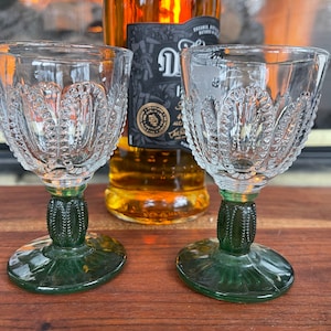 May include: Two clear, vintage-style wine glasses with intricate patterned bowls and green stems. The glasses are displayed on a wooden surface, with a bottle of amber-colored liquor in the background.