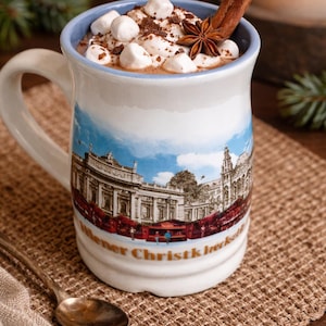 May include: A mug of hot chocolate topped with marshmallows, cocoa powder, a star anise, and a cinnamon stick. The mug has a blue rim and a scenic image of a European city. A spoon rests on a woven mat.