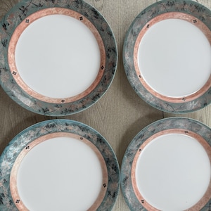 May include: Four white ceramic plates with a decorative border. The border features a teal and peach color scheme with a floral and diamond pattern. The plates are arranged in a square formation on a wooden surface.