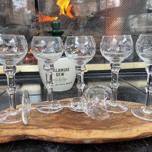 May include: A set of six clear, etched glass cordial glasses with floral designs. Five glasses stand upright, while one lies on its side. The glasses are on a wooden board, with a bottle of Irish whiskey in the background.