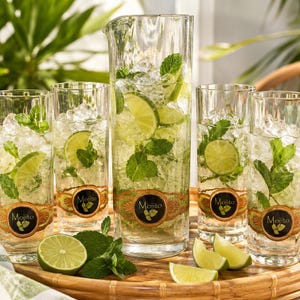 May include: A clear glass pitcher and four matching tall glasses filled with ice, mint leaves, and lime slices. Each piece features a decorative label with the word Mojito and a lime graphic. Fresh lime wedges and mint sprigs sit on the tray.