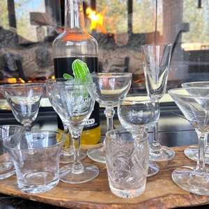 May include: A collection of clear glass drinkware, including shot glasses, stemmed glasses, and martini glasses, arranged on a wooden serving board. A bottle of liquor with a green cactus sticker is in the background. A fireplace is visible in the background.