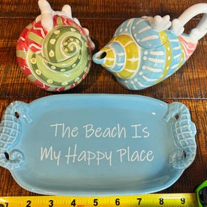 May include: A ceramic seashell teapot and a matching salt shaker, both with colorful designs. A light blue rectangular tray with seahorse handles and the text "The Beach Is My Happy Place" is also included.