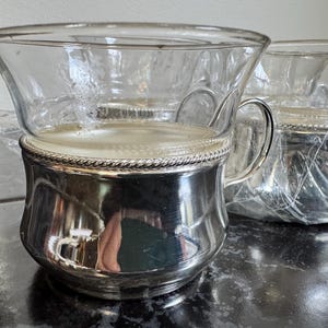 May include: A clear glass cup with a silver-colored metal base and handle. The rim of the metal base has a decorative rope-like design. The cup is sitting on a black surface. Another cup is visible in the background.