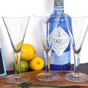May include: Three clear stemmed cocktail glasses, a blue gin bottle, lemons, and a cocktail book are arranged on a wooden board. The gin bottle has a blue label with white text. The book cover has the words "Iconic Cocktails".