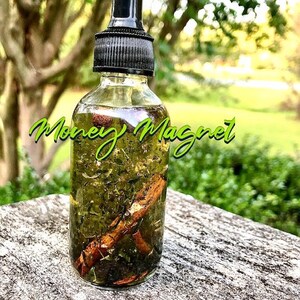 Pode incluir: Uma garrafa de vidro transparente com uma tampa conta-gotas preta, cheia de um l&iacute;quido verde e elementos bot&acirc;nicos. A garrafa tem as palavras "Money Magnet" em verde. A garrafa est&aacute; sobre uma superf&iacute;cie cinza desgastada.