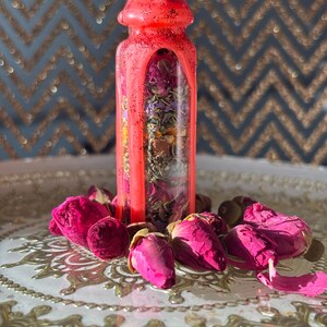 Puede incluir: Una botella de vidrio transparente llena de flores y hierbas secas, sellada con una tapa de cera rosa brillante. La botella est&aacute; rodeada de capullos y p&eacute;talos de rosas rosas sobre una bandeja plateada decorativa.