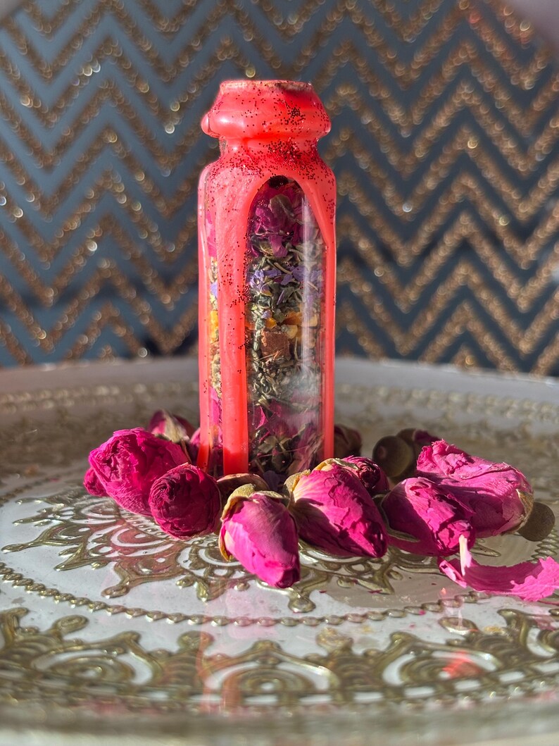 Puede incluir: Una botella de vidrio transparente llena de flores y hierbas secas, sellada con una tapa de cera rosa brillante. La botella est&aacute; rodeada de capullos y p&eacute;talos de rosas rosas sobre una bandeja plateada decorativa.